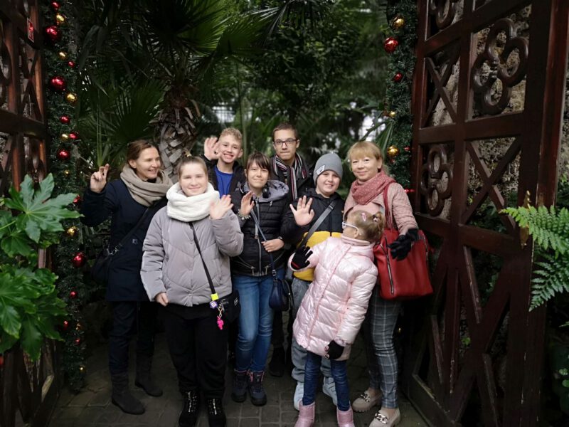 Zdjęcie przedstawia grupę uśmiechniętych dzieci i dorosłych przy wejściu do palmiarni. Wejście ozdobione jest czerwonymi bombkami.W głębi znajduje się gęstwina roślinności.